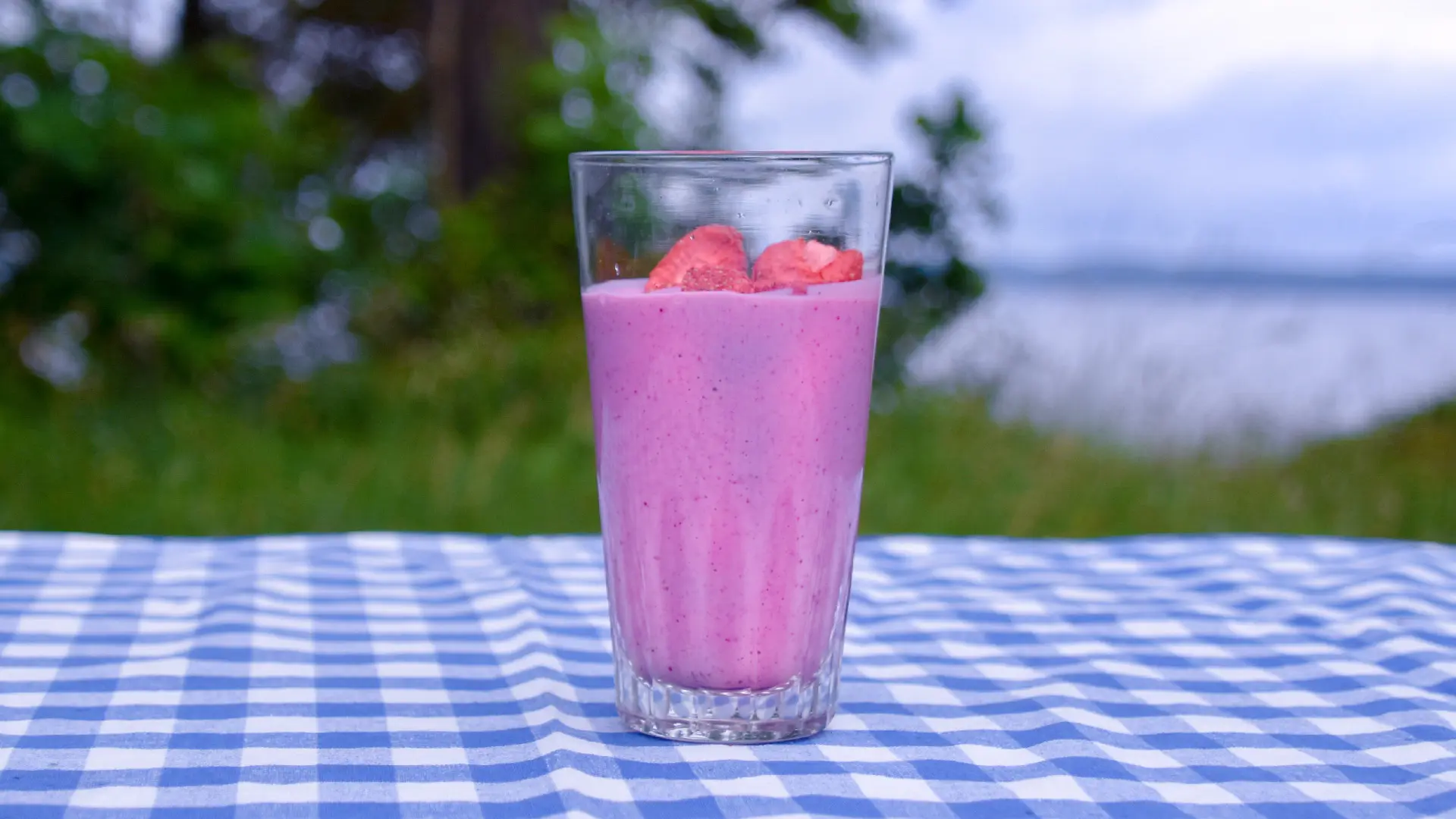Smoothie gjord på frystorkade röda bär, dekorerad med stora frystorkade jordgubbar. Står på ett bord med blåvitrutig duk på ett bord utomhus.